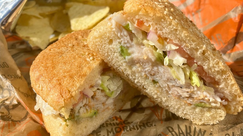 A Schlotzsky's sourdough sandwich cut in half in front of a bag of chips and branded cup