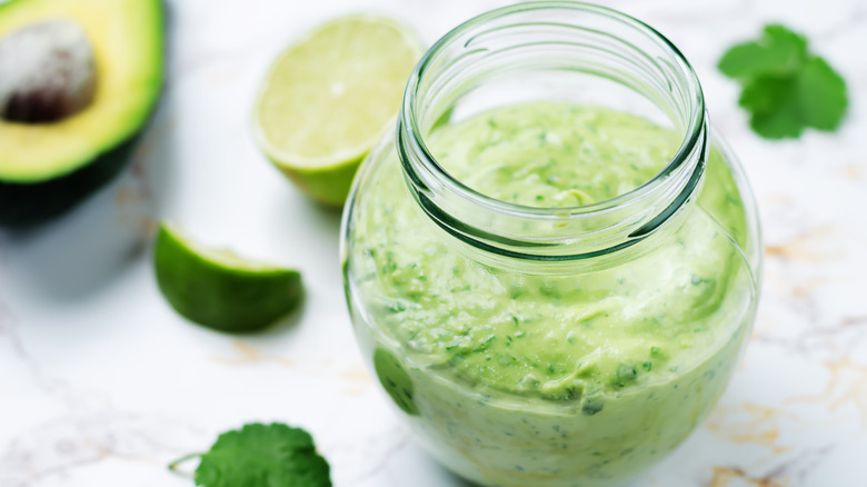 cilantro lime crema in a jar against white backdrop with lime, cilantro, and avocado