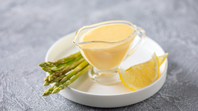hollandaise sauce in a jar on a white plate with asparagus and lemon on a black table