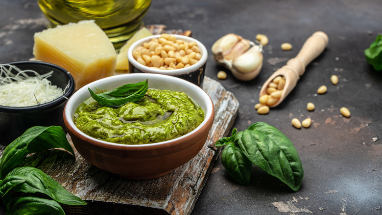 pesto sauce in a jar with spoon against light backdrop with basil, garlic, pine nuts, and cheese kept around it