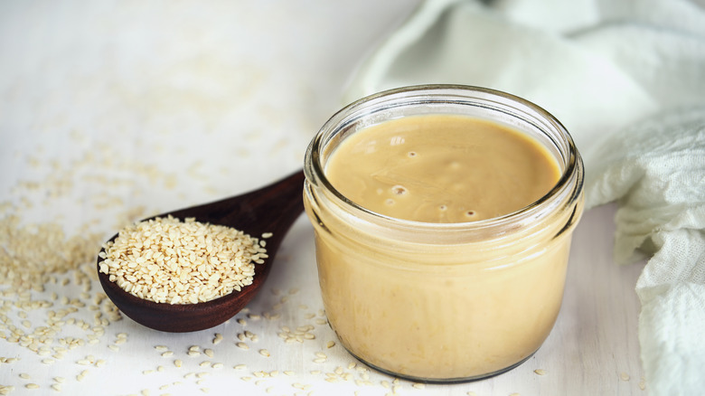 tahini sauce in a jar on a wooden table with sesame seeds, cilantro, and lemon