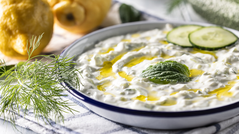 tzatziki sauce in a white bowl on a kitchen towel with mint, cucumber, lemon and dill placed around it