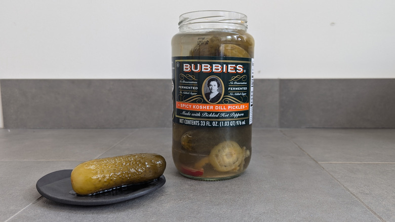 Tall glass jar of Bubbies whole pickles with a whole pickle on a plate