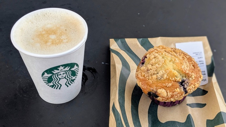A cup of coffee in a paper cup topped with foam next to a blueberry muffin on a black table