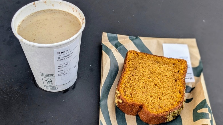 A paper cup of cinnamon coffee with a slice of pumpkin loaf on a black table
