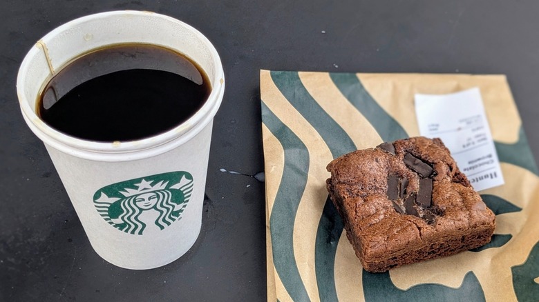 A cup of black coffee in a paper cup next to a chocolate brownie on a black table