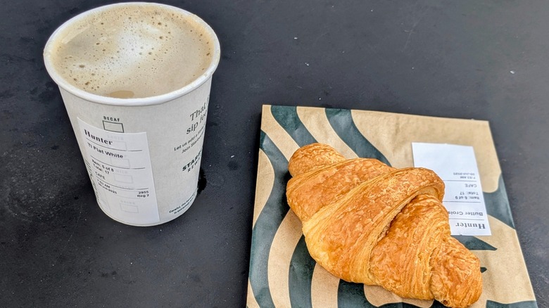 A flat white in a paper cup next to a croissant on a black table