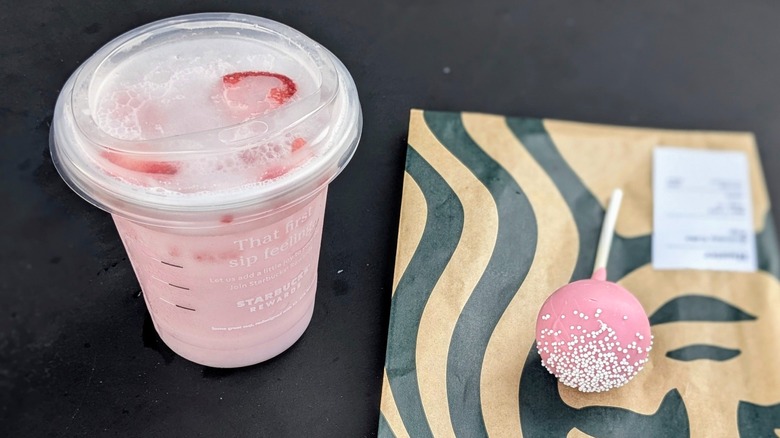 An iced Pink Drink in a plastic cup next to a pink cake pop on a black table