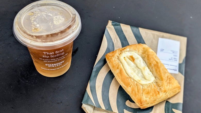 A plastic cup of cold brew next to a cheese danish on a black table