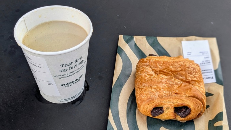 A white chocolate mocha in a paper cup next to a chocolate croissant on a black table