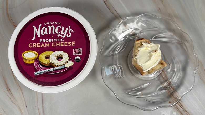 Package of Nancy's cream cheese next to bagel piece with cream cheese on glass plate