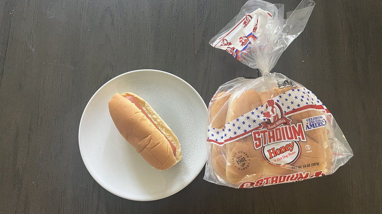 Aunt Millie's Stadium Honey Hot Dog Bun beg next to a hot dog served on a white plate