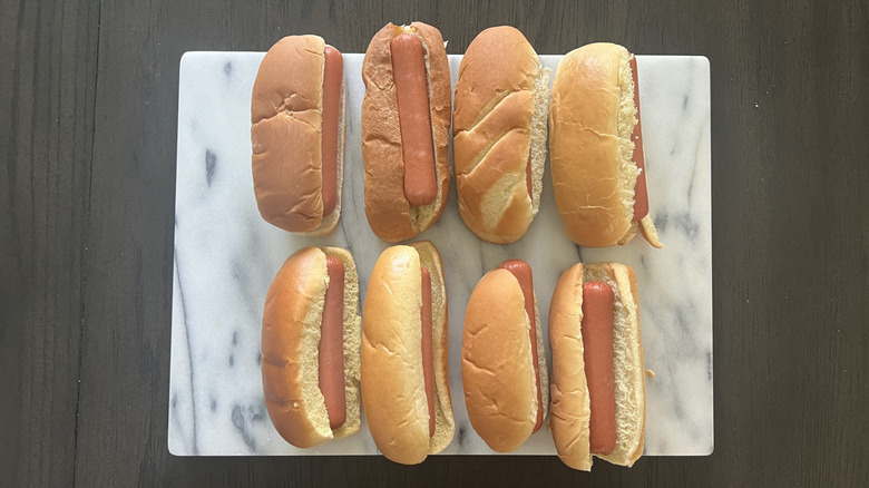 Eight different brands of hot dog buns with hot dogs laid on a marble board on a dark counter