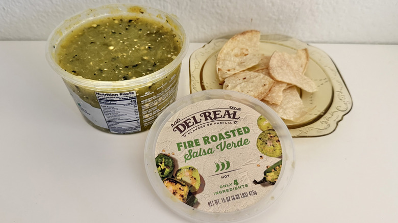 A plastic container of Del Real Fire Roasted Salsa Verde on a table with chips on a plate next to it