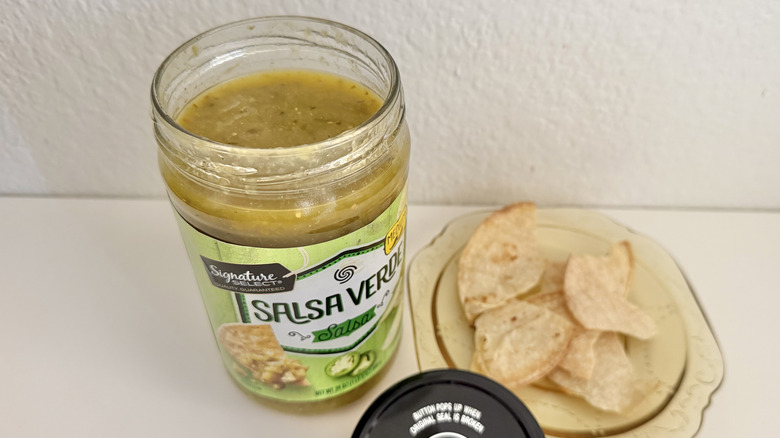 Open jar of Signature Select Salsa Verde next to some tortilla chips on a plate