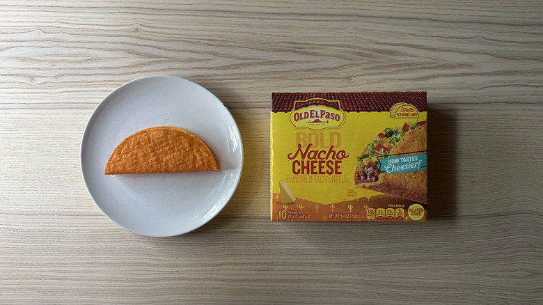 A box of Old El Paso Bold Nacho Cheese Taco Shells on wooden table with one shell on a plate