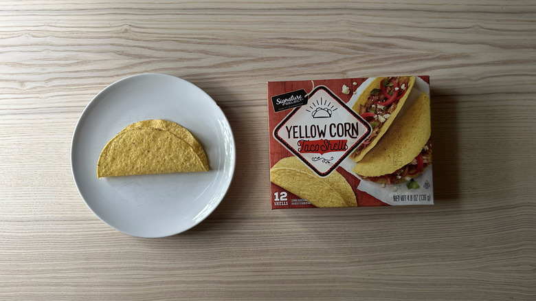 A box of Signature Select Yellow Corn Taco Shells on wooden table with a plate next to it