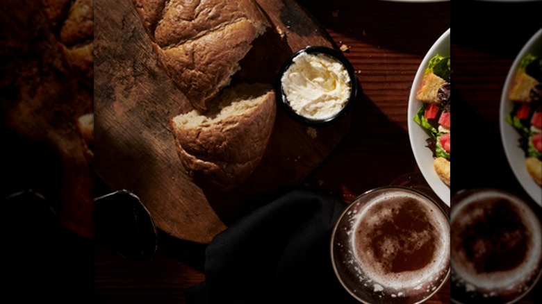 Loaf of honey wheat bread with piece torn off and cup of butter on cutting board.