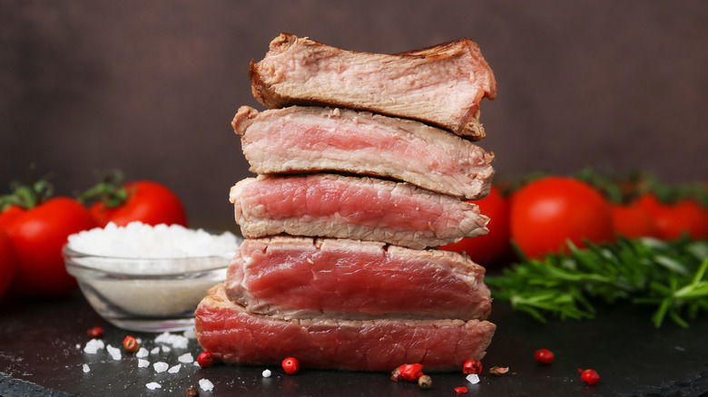 Sliced pieces of cooked beef in varying doneness from well-done to rare with small glass bowl of salt and tomatoes in background.