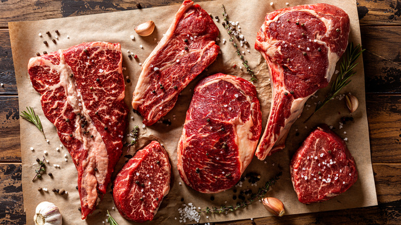 Six different cuts of raw steak with salt and pepper and herbs on brown parchment paper sitting on wood surface.
