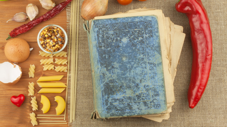 Birds-eye view of a very old blue cookbook placed next to dried pasta, garlic, chile, egg, and onion