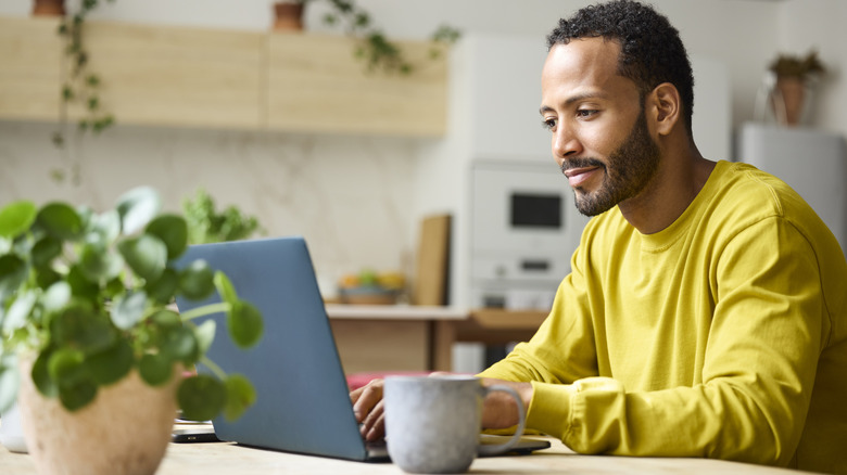 Bearded man in a yellow shirt working at a laptop near his kitchen