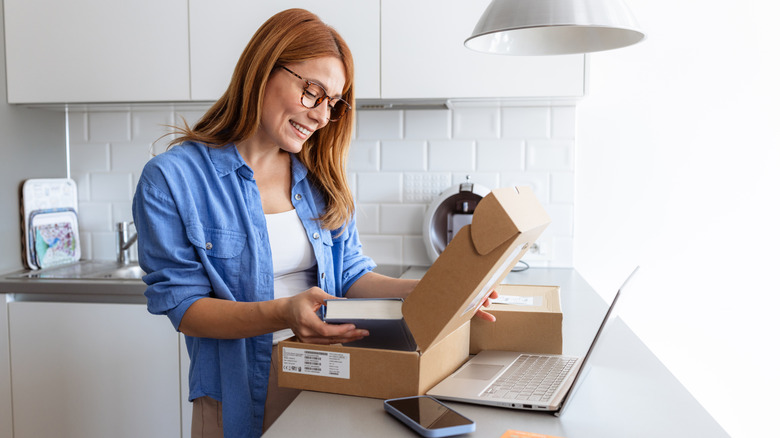 Woman wearing a blue shirt and opening a delivery package containing a book in a kitchen