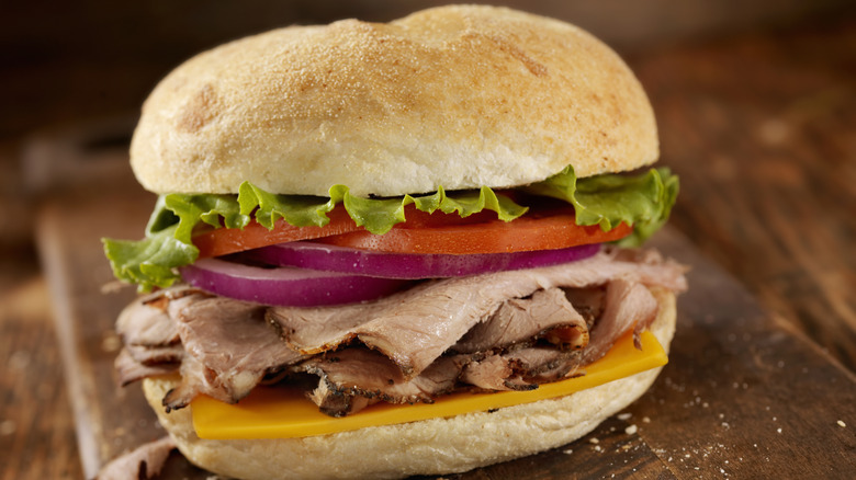 close up of a roast beef sandwich on cutting board with veggies and cheese