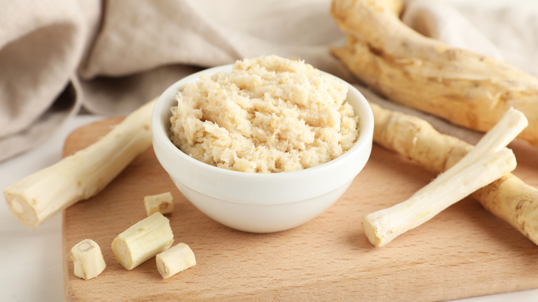 fresh and shredded horseradish on a cutting board surrounded by horseradish roots