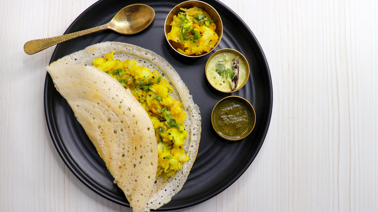 Dosa filled with spiced potatoes, alongside spoon and chutneys on plate