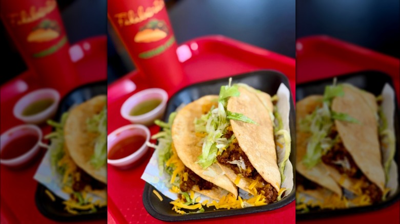 Two tacos on a red tray with red and green sauce and a Filiberto's drink cup