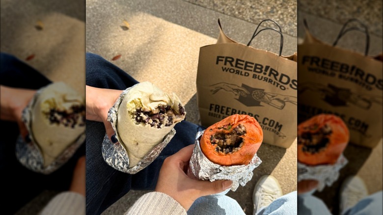 Two people holding burritos with bites taken out next to a Freebirds World Burrito Bag outside