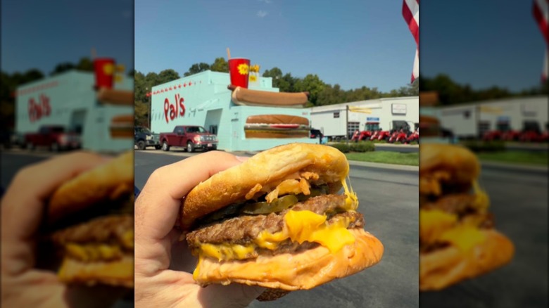 A hand holding a burger with cheese and peppers in front of Pal's Sudden Service