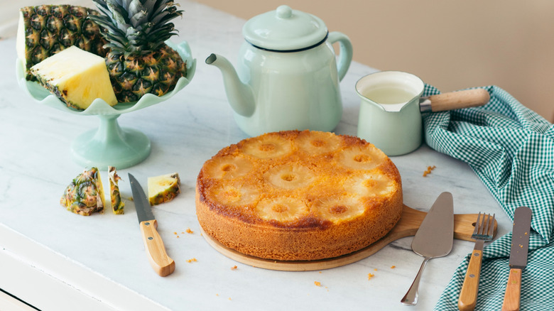 Pineapple upside down cake on wooden platter with kettle and fresh pineapple on table