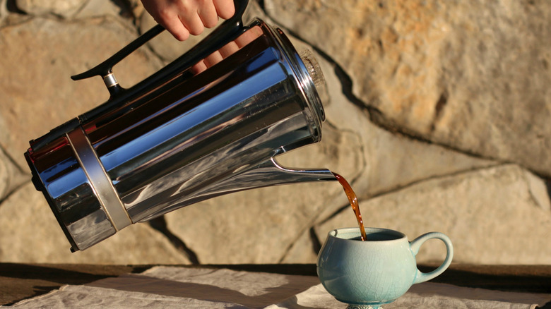 pouring coffee from a vingage electric percolator into a period-matching cup, on a patio table against a stone wall