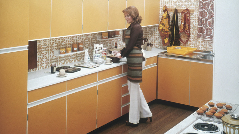A woman standing in a 1970s-style kitchen with minimalist yellow cabinets and an intricate tile backsplash