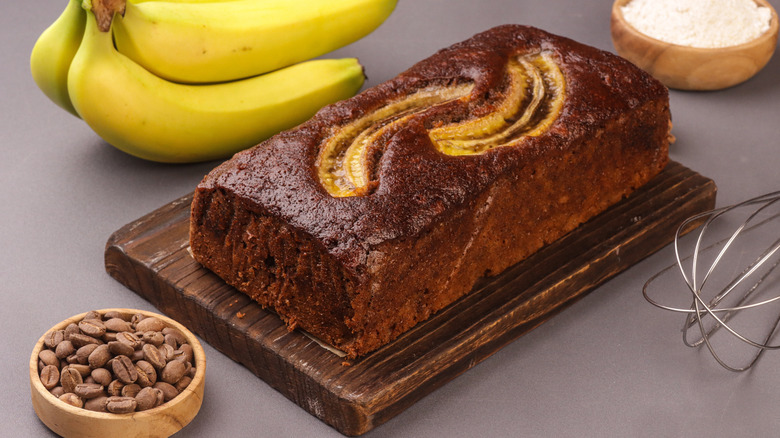 Freshly baked banana bread surrounded by flour, bananas, coffee beans, and whisk