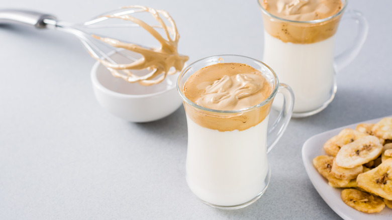 Two glasses of dalgona coffee alongside plate of banana chips and whisk