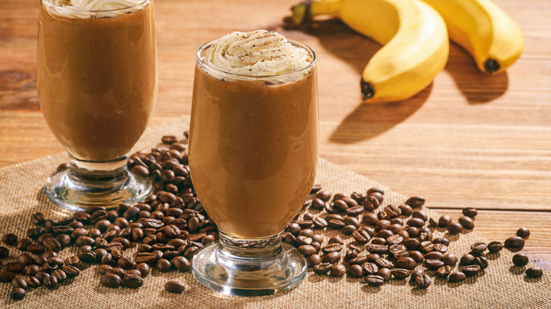 Two glasses full of coffee banana smoothie, topped with whipped cream and surrounded by coffee beans and bananas