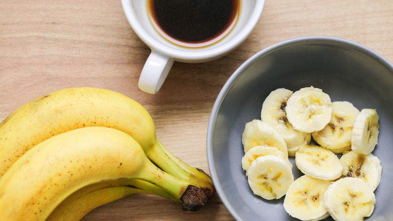 Overhead shot of bananas, some sliced in a bowl, alongside mug of coffee on table