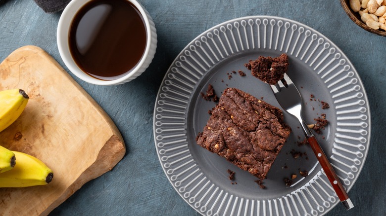 Overhead shot of brownie on plate, alongside cup of coffee, peanuts, and bananas on cutting board