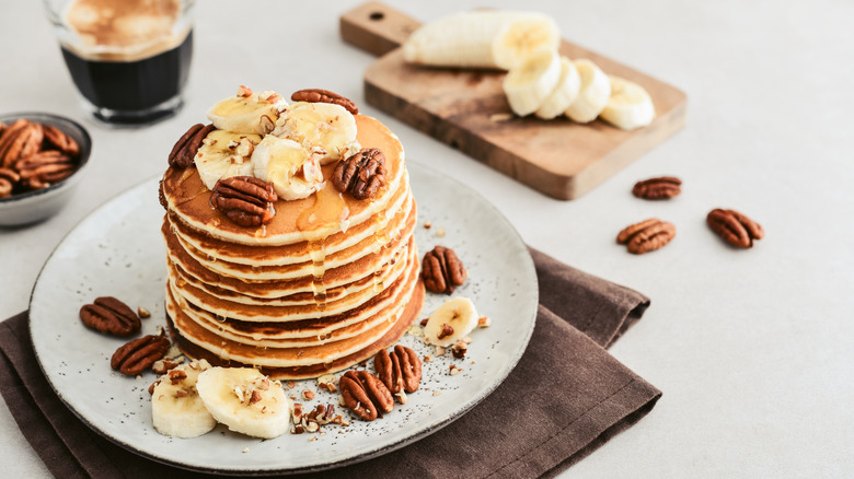 Stack of pancakes topped with bananas, pecans, and maple syrup, with glass of coffee in background