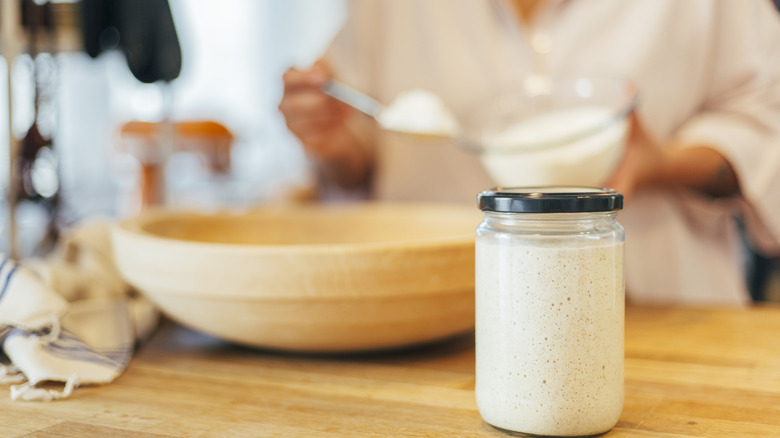 Sourdough starter in jar with lid on counter, woman baking behind