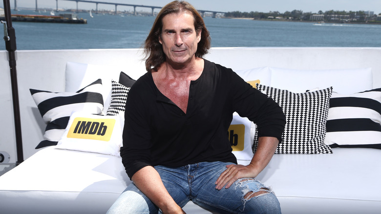 Model Fabio sits on a padded white bench in a black V-neck shirt near the water.