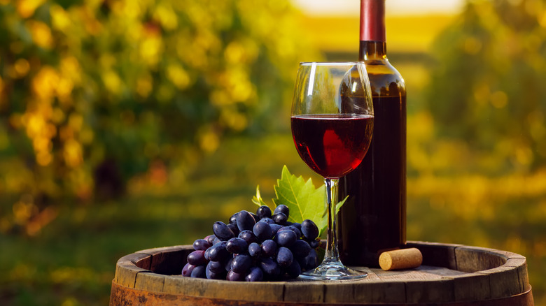 An uncorked bottle of red wine, a glass of red wine, and a bunch of purple grapes rest on a wooden barrel.