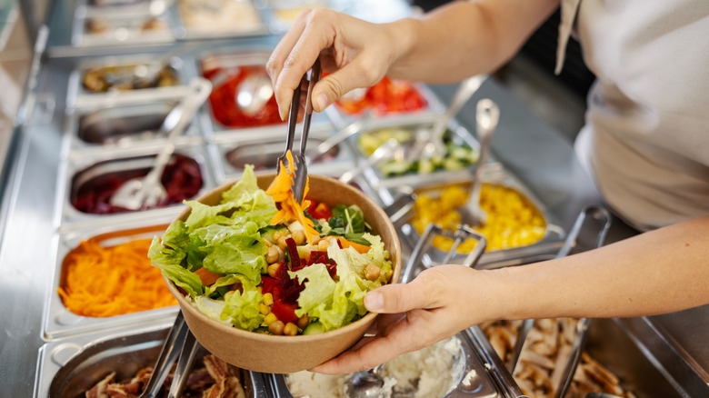 A woman building a large salad in a bowl while visiting a salad bar