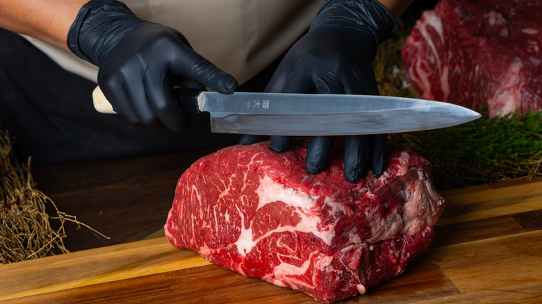 A butcher wearing black rubber gloves holding a knife and slicing a piece of meat