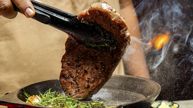 A piece of steak in tongs being seared on a cast iron skillet