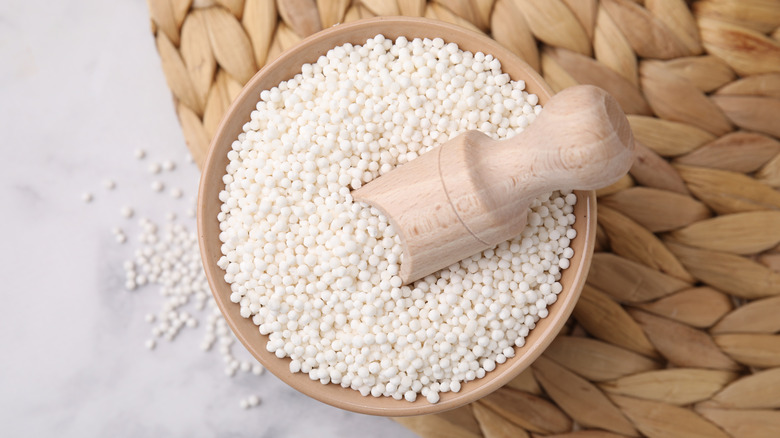 White dried tapioca pearls in a wooden bowl with a wooden scoop against a straw mat background.