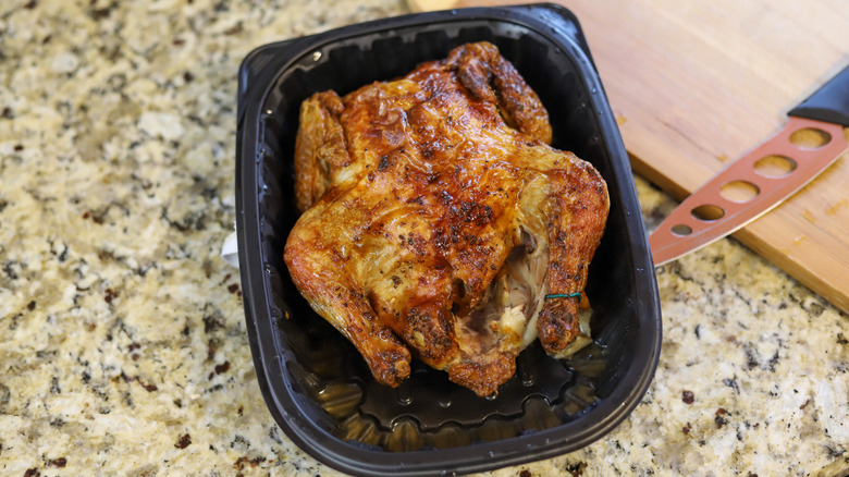 A rotisserie chicken in a black plastic container sitting on a kitchen counter next to a knife